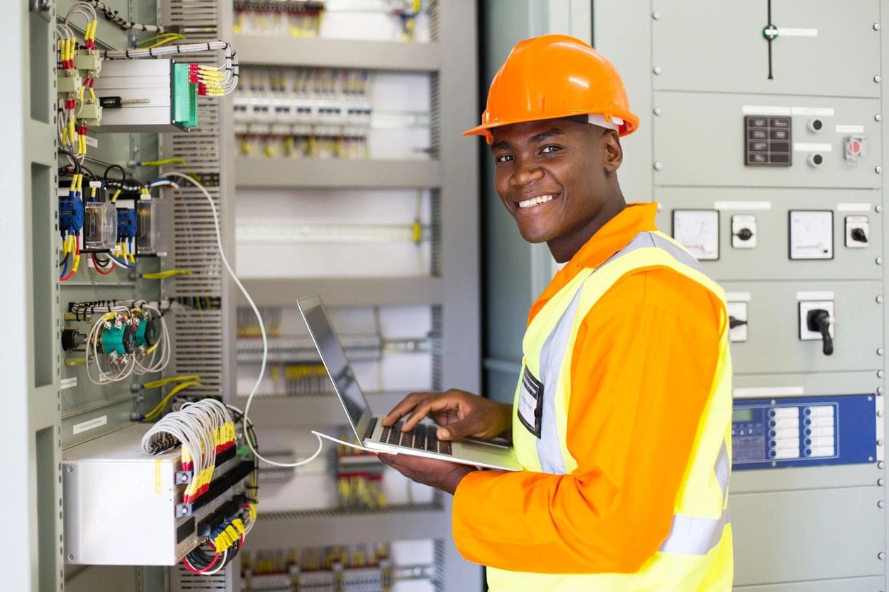 A man in an orange hard hat and safety vest holding a laptop.